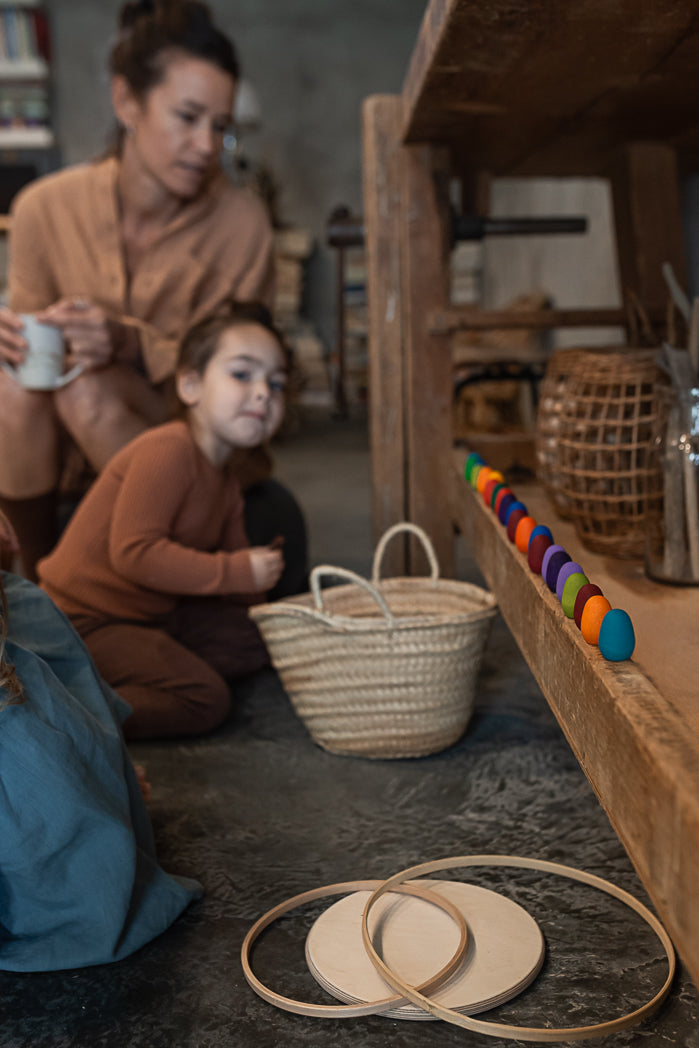 a child and her parent sit in a room with a woven basket. the mandala rainbow wooden eggs are lined up along a ledge