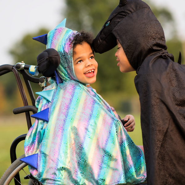two children one standing and one in a wheelchair are laughing with their dragon cape costumes on